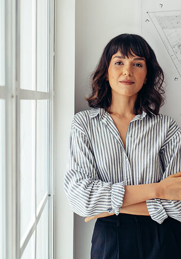 Portrait of female architect standing beside architecture drawings in office. Businesswoman standing in office with arms crossed.